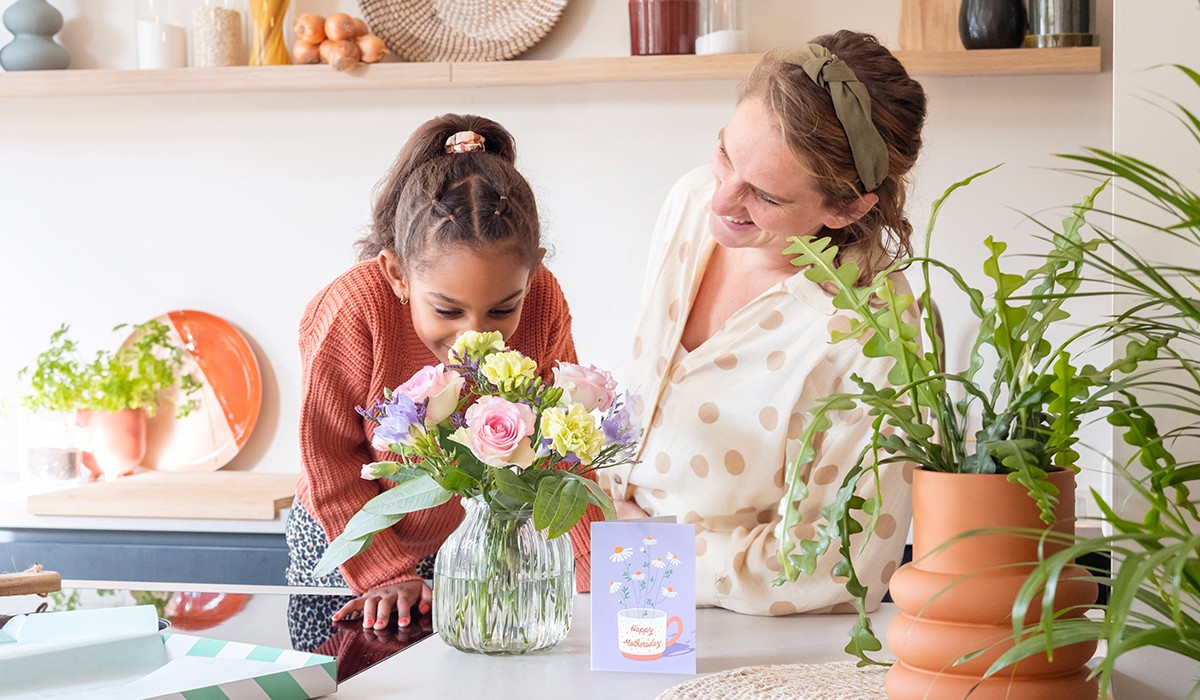 Meisje ruikt aan een bos bloemen met daarnaast een moederdagkaart. Moeder kijkt er lachend naar. 